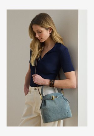 Kvinde med langt blondt hår iført navy blå ribbet trøje og beige bukser, holder en lyseblå håndtaske med guldlås, stående op ad en væg.