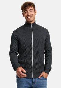 Suéter de cremallera color gris oscuro con un patrón de espiga, cuello alto, puños y dobladillo acanalados, combinado con jeans de color azul claro.