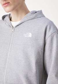 Sweatshirt à fermeture éclair gris en tissu doux, doté d'une poche avant, avec un logo blanc sur la poitrine gauche et une capuche à cordon de serrage ajustable.