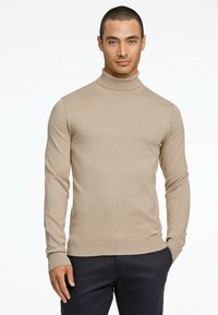 Suéter de cuello alto beige hecho de tejido de punto texturizado, con mangas largas y un diseño ajustado. Llevado con pantalones oscuros.