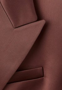 Hnedý blazer s jemnou textúrou, ostro tvarovaným klopom a vreckom s paspulou. Materiál sa javí ako ľahký s elegantným povrchom.