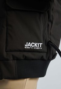 Sort polstret jakke med ribbede manchetter, der har hvid broderet tekst "JACK1T" og koordinater. Inkluderer en sidelomme.