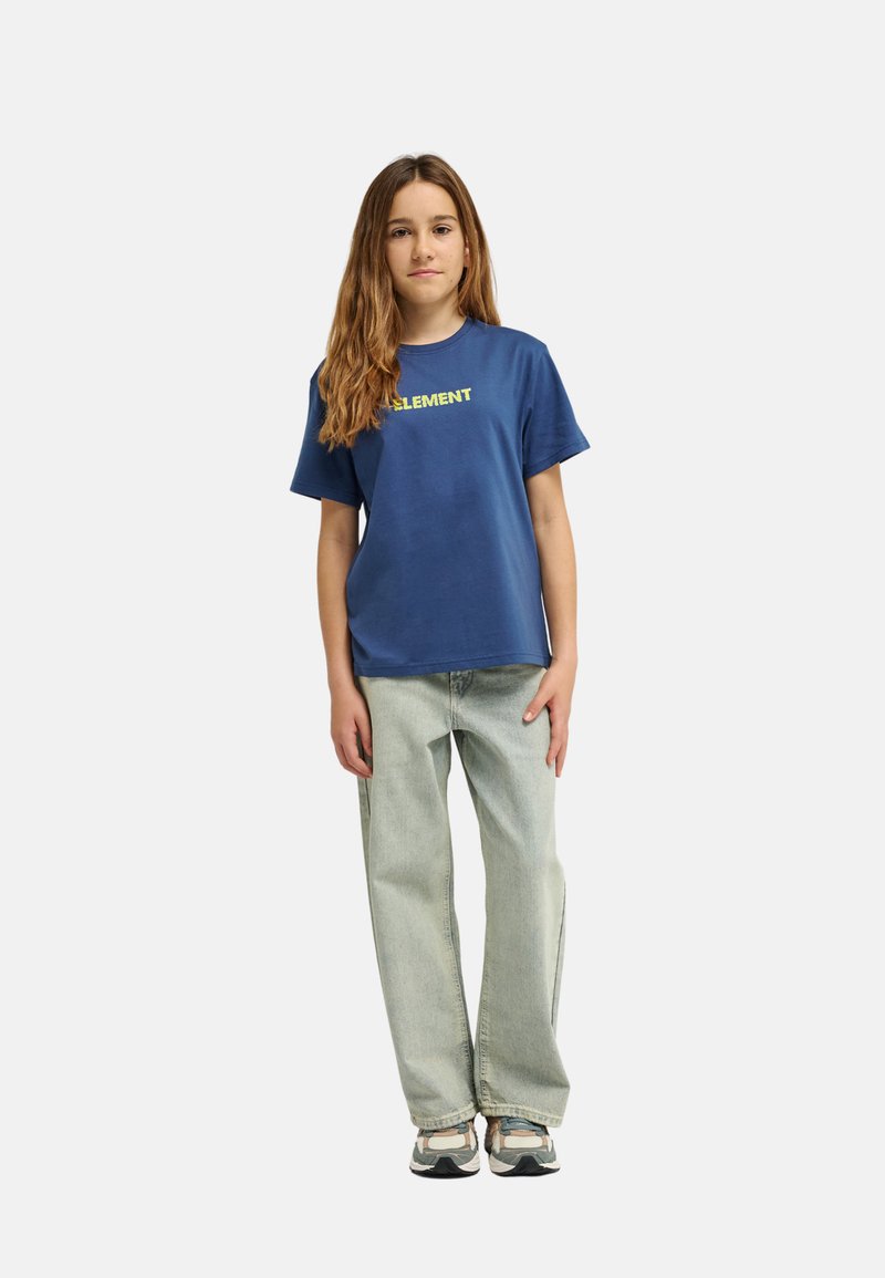 Niña con cabello largo que lleva una camiseta azul con la palabra "ELEMENT", jeans gris claro y zapatillas multicolores, de pie sobre un fondo blanco.