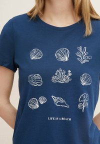 Marinblå T-shirt med vita linjeteckningar av snäckor och koraller, med texten "LIVET ÄR EN STRAND" centrerad längst ner.