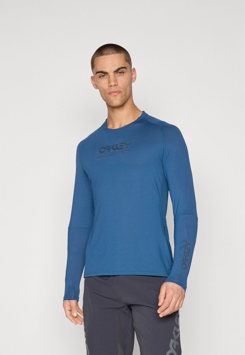Camisa azul de manga larga hecha de tejido transpirable. Presenta un cuello redondo y un logo negro de "Oakley Factory Pilot" en el pecho.