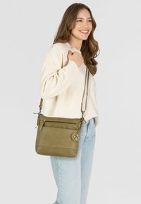 Bolso cruzado de cuero verde oliva con herrajes plateados, correa ajustable, bolsillo delantero con cremallera y un charm de ancla.