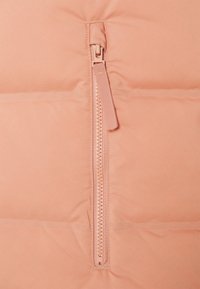 Veste matelassée rose clair avec une fermeture éclair à l'avant et un tirant en tissu. La surface a une texture lisse et mate avec des sections matelassées.