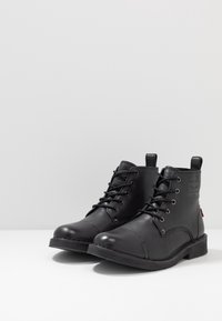 Botas de cuero negro hasta el tobillo con cordones en la parte delantera, punta redondeada, superficie texturizada y suela de goma plana; cuenta con un logo en el lateral y una pestaña en la parte trasera.