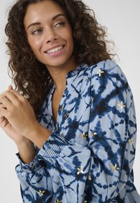 Modrá tie-dye blůza s volánkovým výstřihem, nařasenými manžetami a zlatými květinovými akcenty. Materiál je lehký a vzdušný.