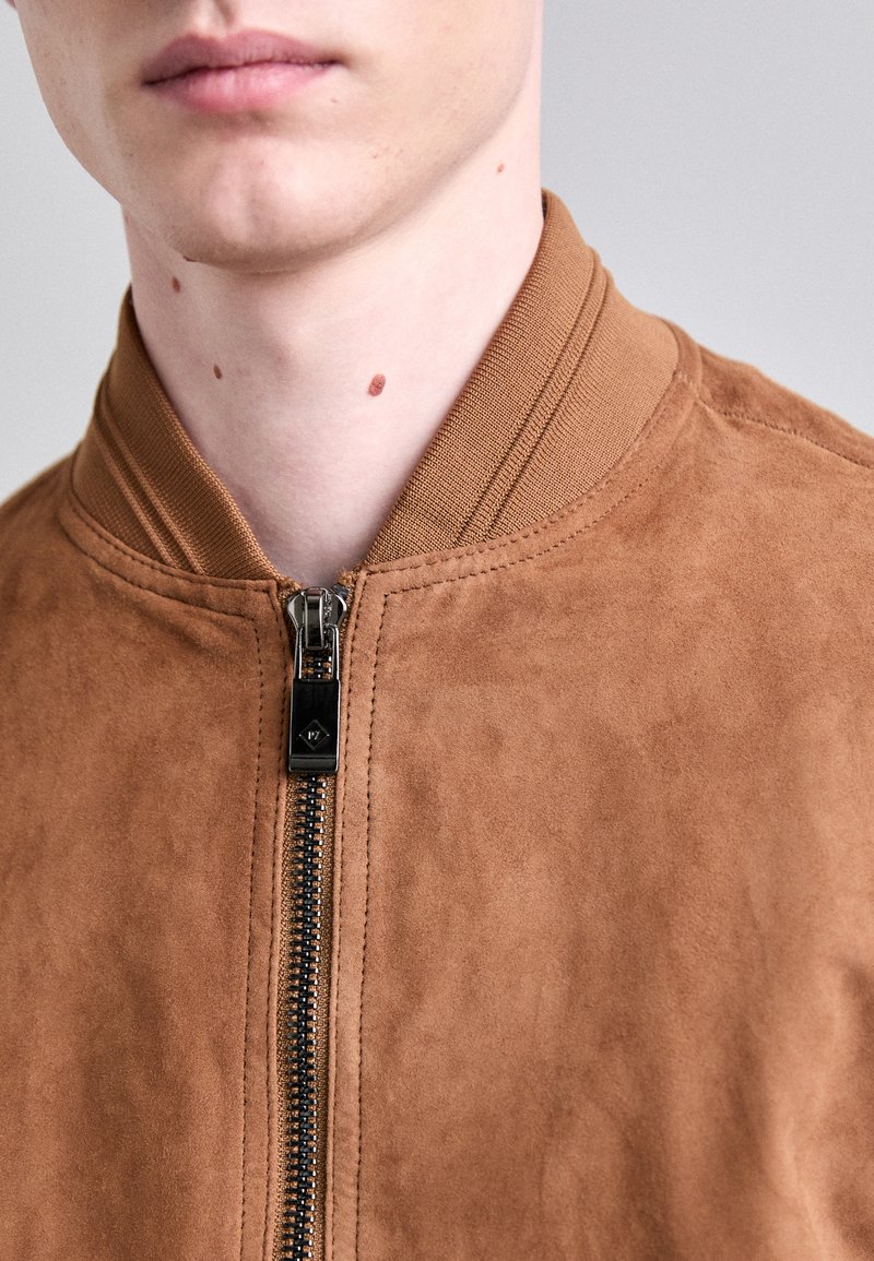 Chaqueta bomber de ante marrón con cierre frontal de cremallera y cuello acanalado. La textura es suave, con detalles de costura mínimos.