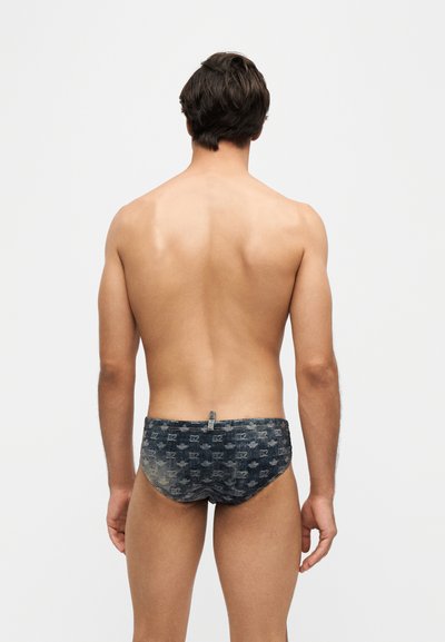 Homme aux cheveux courts et foncés portant un slip de bain foncé à motifs, vu de dos sur un fond clair uni.