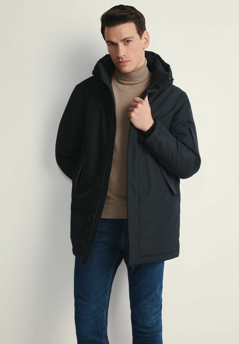 Ciemnonoieska parka z kapturem, zapinana na zamek, z dwoma bocznymi kieszeniami. Nosi się ją na beżowym golfie i niebieskich dżinsach. Gładka faktura.