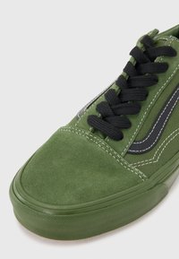 Zapatillas de ante verde con cordones negros, que cuentan con una suela de goma texturizada y detalles de costura blanca. Diseño clásico con acentos de franjas laterales.