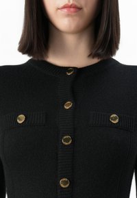 Donna che indossa un cardigan lavorato a maglia nero con bottoni rotondi dorati lungo il davanti e su due taschini sul petto, con capelli corti e lisci di colore scuro.