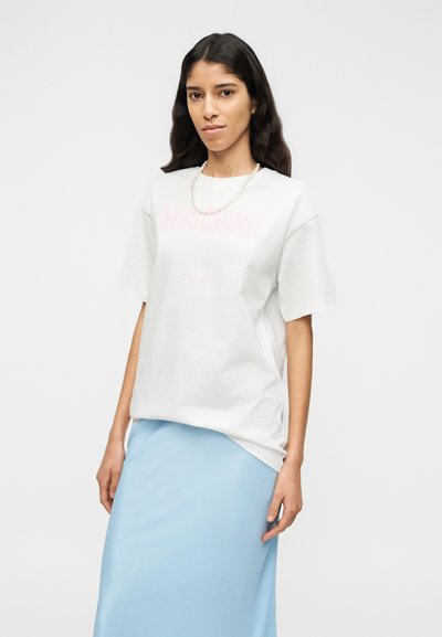 Άσπρο βαμβακερό μπλουζάκι με ροζ λογότυπο Moschino, σε συνδυασμό με ανοιχτό μπλε φούστα. Το μοντέλο φοράει κολιέ με πέρλες, με ομαλές υφές.