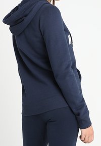 Marinblå hoodie, bomullsblandning, har en känguruficka, ribbade ärmslut och en dragsko i huva. Baksidan har en vit logotyp.