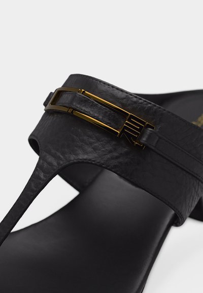 Κοντινό πλάνο του λουριού δερμάτινου σανδαλιού σε μαύρο χρώμα με υφασμένη υφή και λεπτομέρεια χρυσής ορθογώνιας αγκράφας.