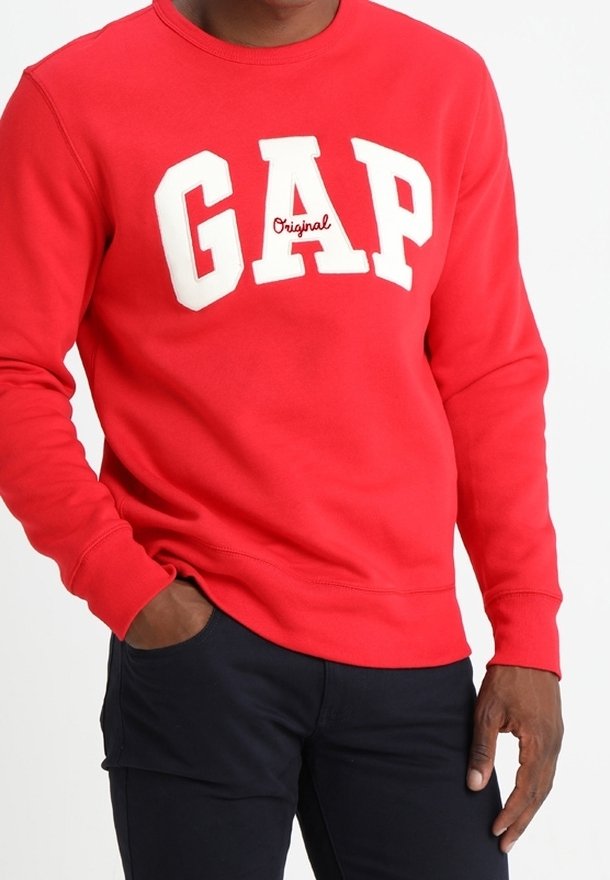 Homme portant un sweat-shirt rouge avec l'inscription blanche "GAP Original", une main dans la poche et un pantalon foncé.