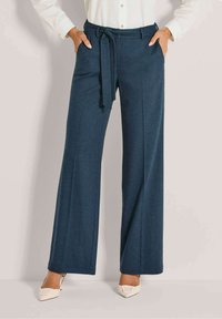 Vrouw in hoge, wijde, marineblauwe broek met een bijpassende stoffen riem, gecombineerd met een witte blouse en witte puntige schoenen.