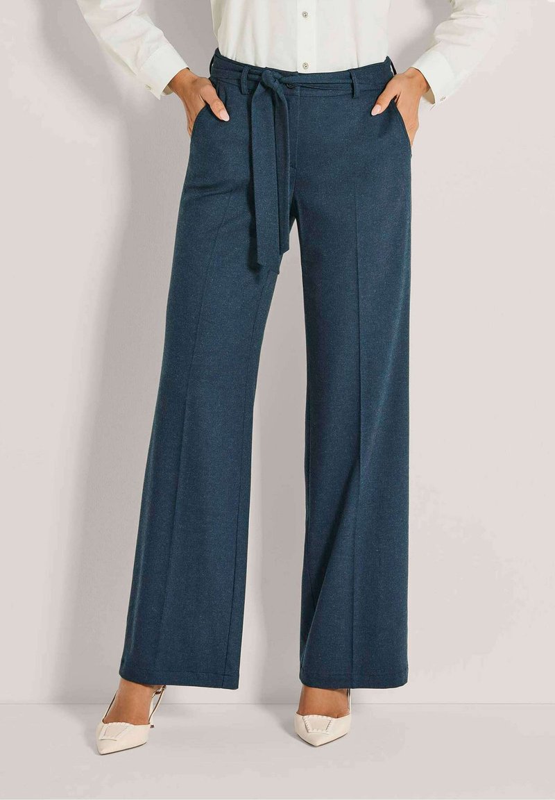 Vrouw in hoge, wijde, marineblauwe broek met een bijpassende stoffen riem, gecombineerd met een witte blouse en witte puntige schoenen.