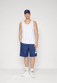 Biely tielko, modré šortky s veľkým bielym logom Nike a biele tenisky. Doplnky tvoria modrá čiapka Nike. Celkový atletický outfit.