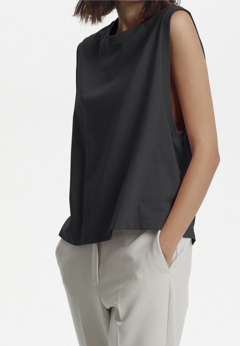 Top senza maniche nero realizzato in tessuto liscio con vestibilità rilassata, caratterizzato da aperture laterali e scollo a girocollo. Abbinato a pantaloni chiari.