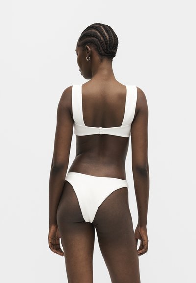 Femme aux cheveux tressés portant un maillot de bain deux pièces blanc, posant dos à la caméra sur un fond uni.