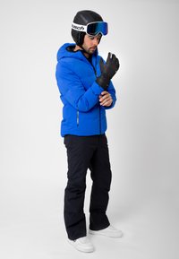 Sininen eristetty hiihtotakki hupulla, mustat käsineet, musta hiihtokypärä ja laskettelulasit. Mustat hiihtohousut ja valkoiset lenkkarit täydentävät asustuksen.