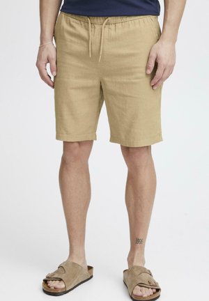Uomo che indossa pantaloncini beige con coulisse e sandali in suede abbinati, in piedi con un tatuaggio visibile "1996" sulla caviglia destra.