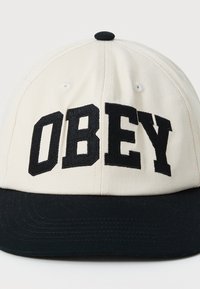 Casquette de baseball avec une couronne en coton beige et une visière noire. Elle présente un lettrage "OBEY" en noir audacieux et des accents noirs contrastants.