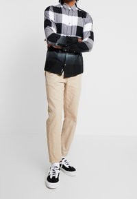 Schwarz-weiß-grau kariertes Hemd mit Farbverlauf, kombiniert mit beigen Hosen und schwarzen Sneakers mit weißen Akzenten.