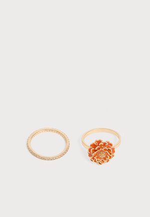 Deux bagues en or sur fond clair, l'une avec de petites pierres claires, l'autre avec un motif en forme de fleur orange.
