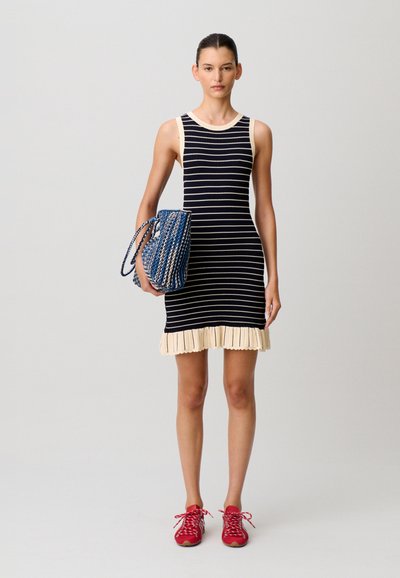 Frau trägt ein ärmelloses, schwarz-weiß gestreiftes Kleid mit einem cremefarbenen Volant am Saum, hält eine blaue geflochtene Tasche und trägt rote Turnschuhe.