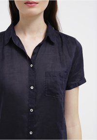 Námořnická modrá košile s krátkým rukávem z lehké tkaniny, s knoflíkovým zapínáním vpředu, kapsou na levé straně hrudi a límečkem.