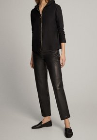 Zwarte rits hoodie met capuchon, gecombineerd met strakke zwarte leren broek en zwarte instapschoenen, waardoor een monotone look ontstaat.