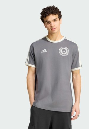 Młody mężczyzna w szarej koszulce Adidas z białymi paskami na rękawach i logo Mercedes-Benz na piersi, stoi z jedną ręką w kieszeni.