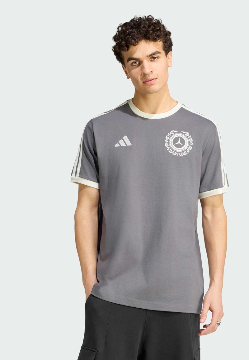 Jeune homme portant un T-shirt gris Adidas avec des bandes blanches sur les manches et un logo Mercedes-Benz sur la poitrine, debout avec une main dans la poche.