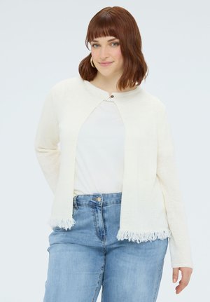 Kvinde med skulderlangt kastanjebrunt hår iført guldringøreringe, en hvid cardigan med frynser, hvid top og højtaljede blå jeans, smilende blidt.