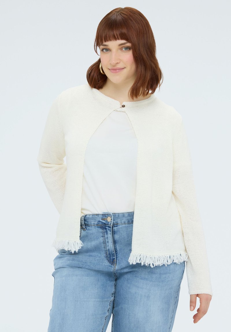 Kvinde med skulderlangt kastanjebrunt hår iført guldringøreringe, en hvid cardigan med frynser, hvid top og højtaljede blå jeans, smilende blidt.