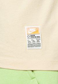 Beżowa koszulka z metką materiałową, z pomarańczowym i czarnym tekstem oraz grafikami, w połączeniu z zielonymi spodniami.