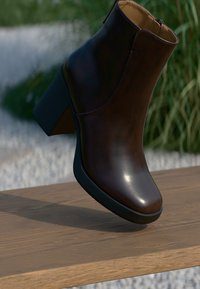 Bota ankle de couro castanho-escuro com salto robusto e fecho de correr lateral, repousando sobre uma superfície de madeira com um fundo de vegetação desfocada.