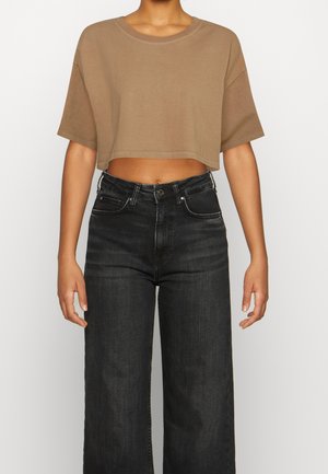 Persoon die een cropped bruine t-shirt en hoge taille zwarte wijd uitlopende jeans draagt, staand tegen een effen achtergrond.