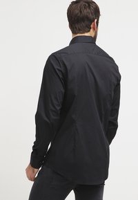 Hombre con camisa negra de manga larga y jeans negros, de espaldas contra un fondo blanco liso.