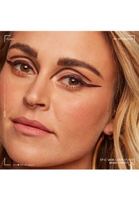 Närbild av ett ansikte med gyllene örhängen, varm hudton, kraftigt brunt eyeliner och nude läppar; texturerad hud och definierade ögonbryn synliga.