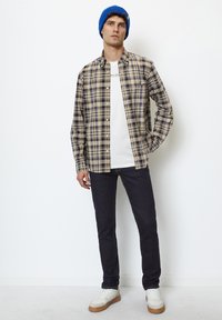 Plaid button-up shirt in beige and navy, layered over a white T-shirt. Slim dark jeans and white sneakers with tan accents. Blue knit beanie.