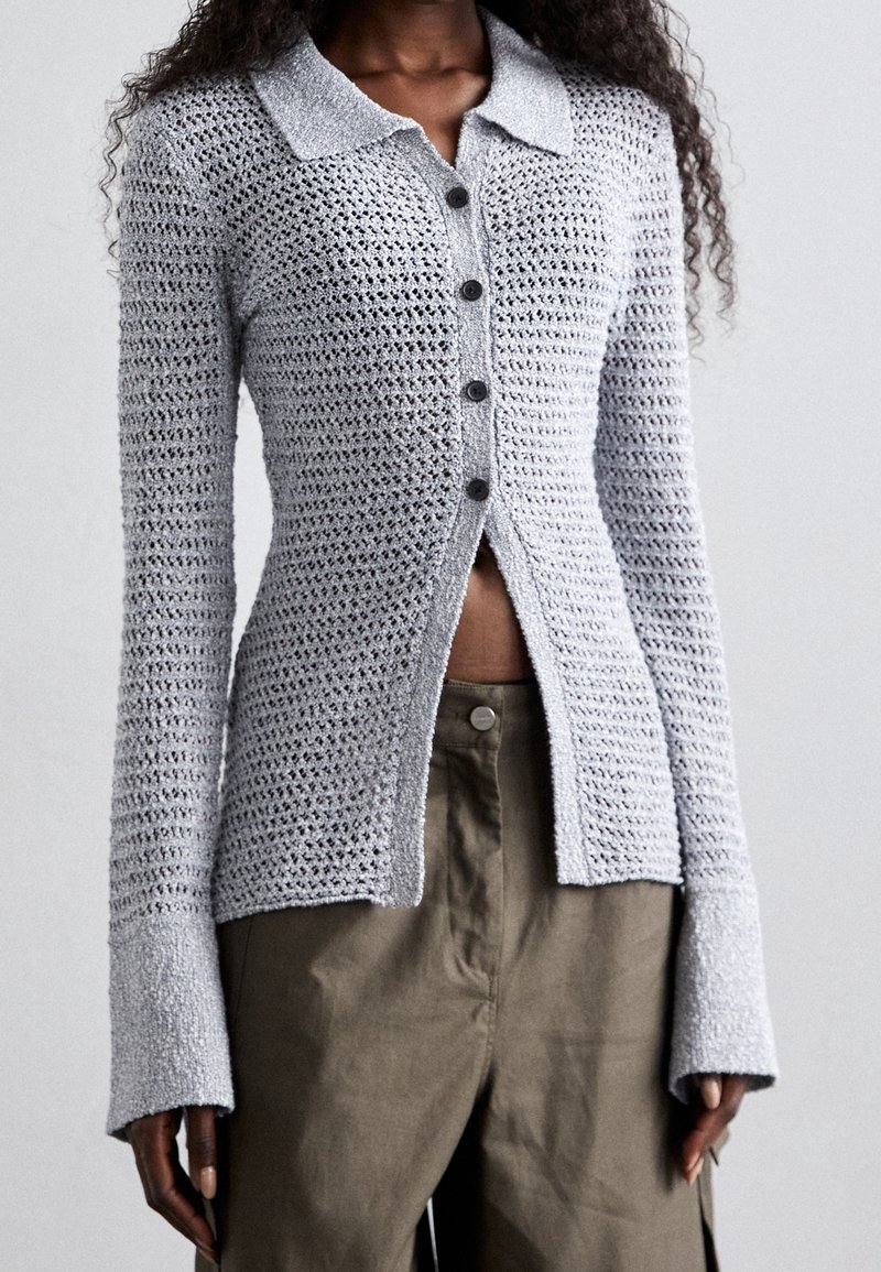 Cardigan lavorato a maglia grigio chiaro con colletto, bottoni frontali e maniche svasate, caratterizzato da un motivo a maglia aperta e una lunghezza leggermente cropped.