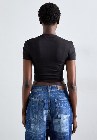 Person with short curly hair stands facing away, wearing a fitted black crop top and high-waisted blue jeans with newspaper print.