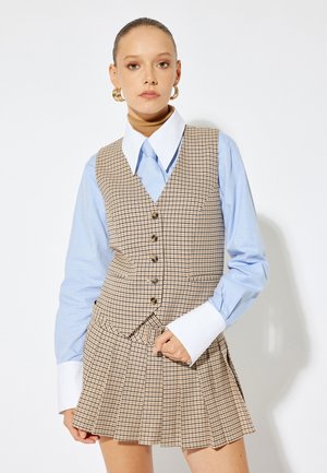 Bruin geruite gilet over een lichtblauw shirt met grote witte manchetten, gecombineerd met een geplooide geruite rok. Gouden knoopaccenten op het gilet.