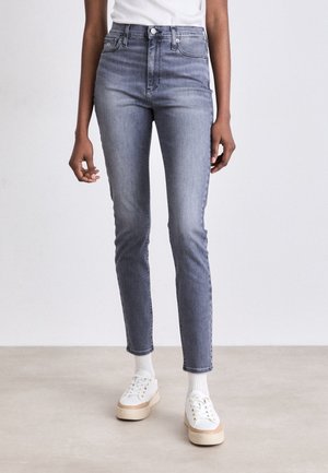 Schmal geschnittene graue Jeans, getragen mit weißen Socken und weißen Plateausneakern, kombiniert mit einem weißen Oberteil an einer stehenden Person.