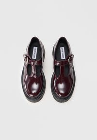 Un par de zapatos Mary Jane de un brillante burdeos oscuro con suelas negras gruesas y correas con hebilla sobre fondo blanco.